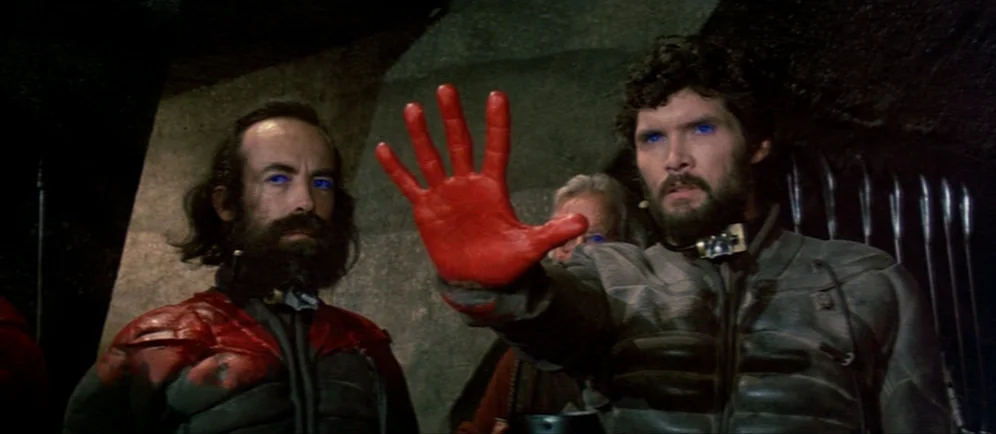 Dune (1984) movie still: medium — Dark rocky interior walls background the two men wearing textured futuristic tactical suits. One man extends his…