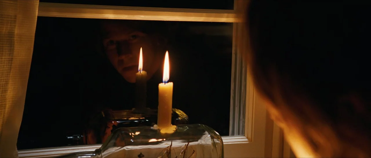 The Lovely Bones (2009) movie still: medium — A dark night scene shows a reflection of a man in a glass window pane. A lit candle sits on a clear…