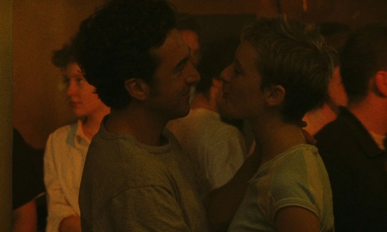 Late August, Early September (1998) movie still: medium — A dimly lit, crowded space filled with people in a low-visibility environment. A man and a…