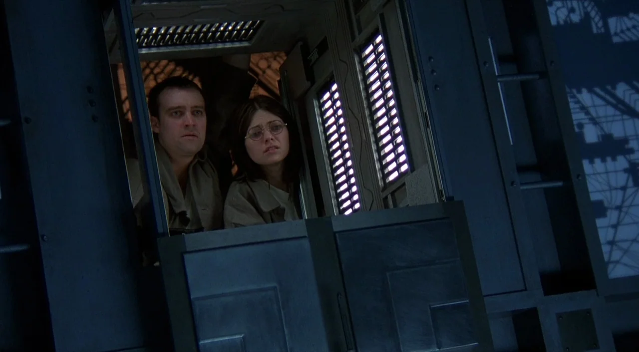 Cube (1997) movie still: medium — The interior of a metallic, dark-toned train car features heavy paneling and narrow windows. A man and a woman…