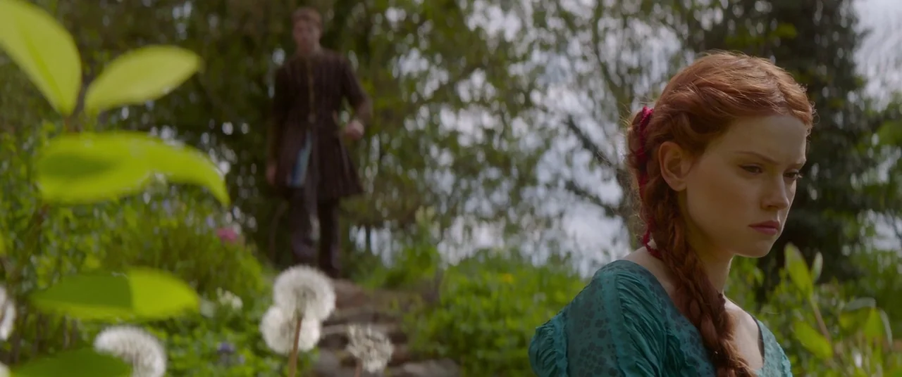 Ophelia (2018) movie still: medium — A lush green garden filled with trees and dandelion seed heads. A woman stands in the foreground while a man…