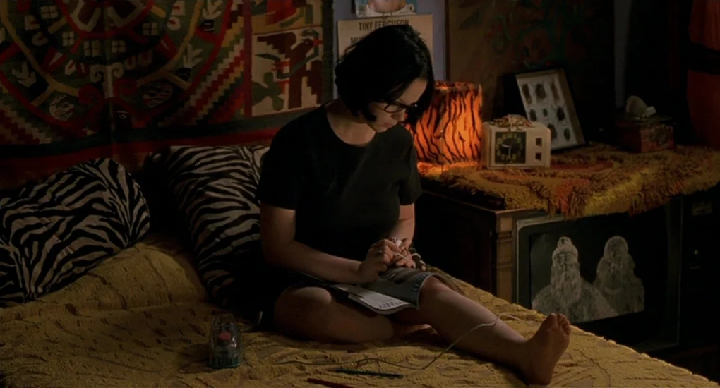 Ghost World (2001) movie still: medium — A dimly lit room with patterned wall hangings and vintage furniture including an old television. A teenager…