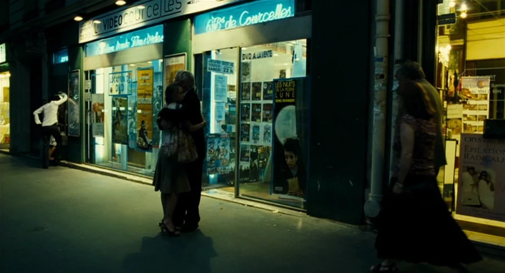 Paris, Je T’Aime (2006) movie still: wide — A Parisian street at night features a neon-lit store front with film posters. A man and woman embrace in…