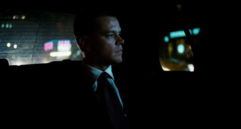 The Adjustment Bureau (2013) movie still: medium — A man sits in the back of a car at night. Blurry city lights glow through the rear window of the…