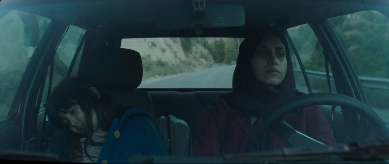 Under The Shadow (2016) movie still: medium — The interior of a moving car showing a driver and a sleeping child passenger. A winding mountain road…
