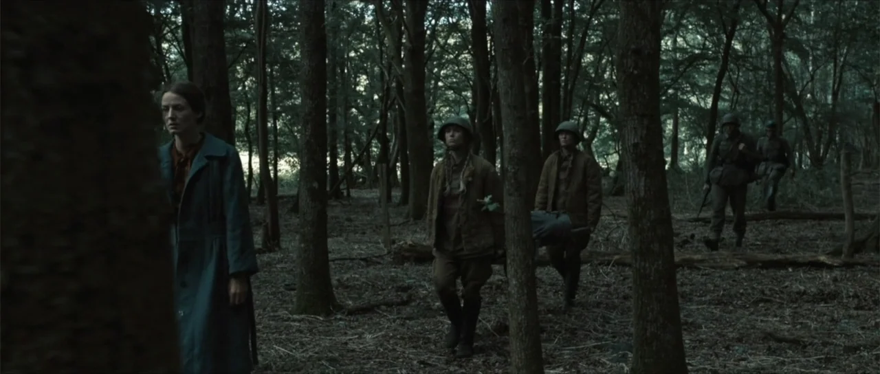 Sin retorno (2009) movie still: fullbody — A dense, shaded woodland filled with tall thin trees and a leaf-strewn ground. A woman in a blue coat…