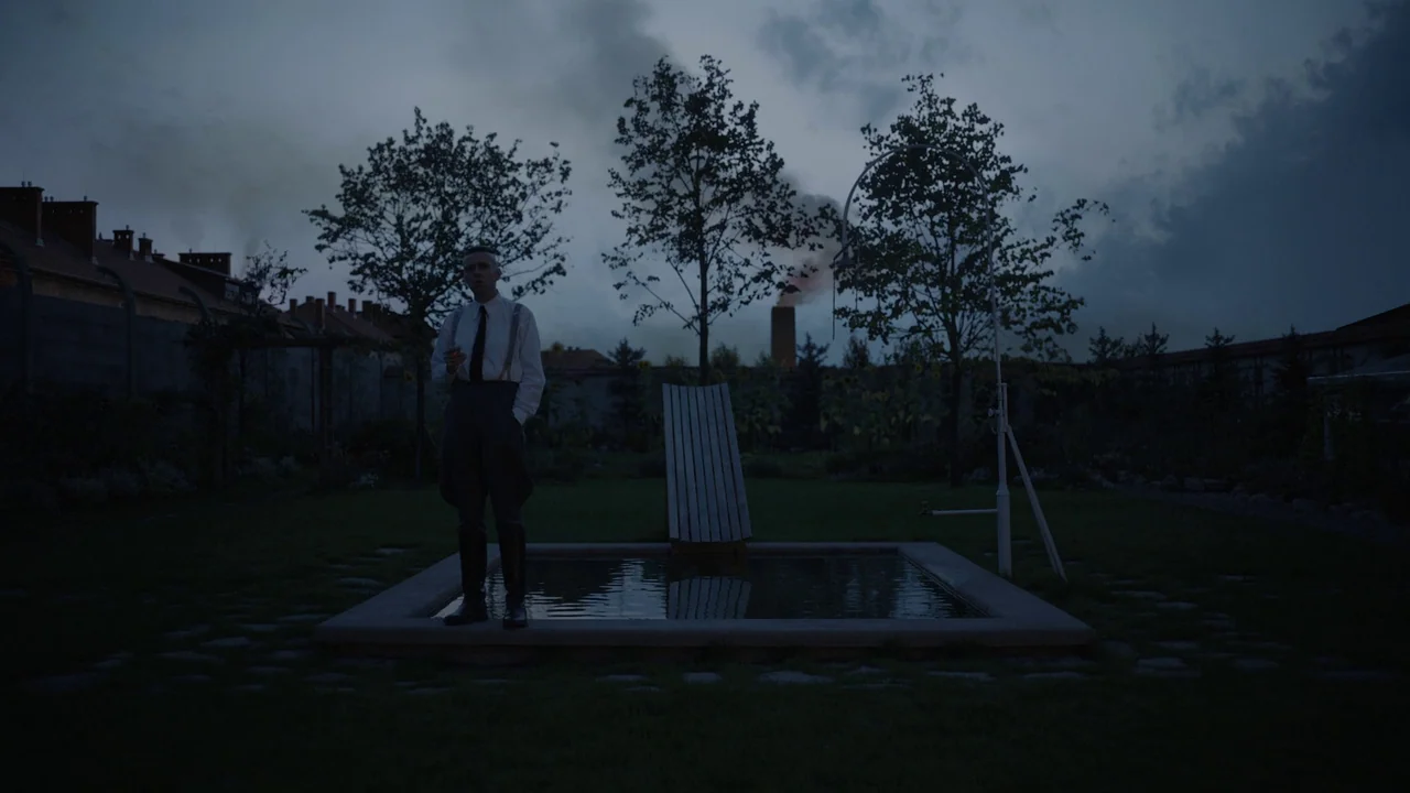 The Zone Of Interest (2023) movie still: fullbody — A dark, overcast backyard features a small stone-edged pool and sparse trees. A man in formal…