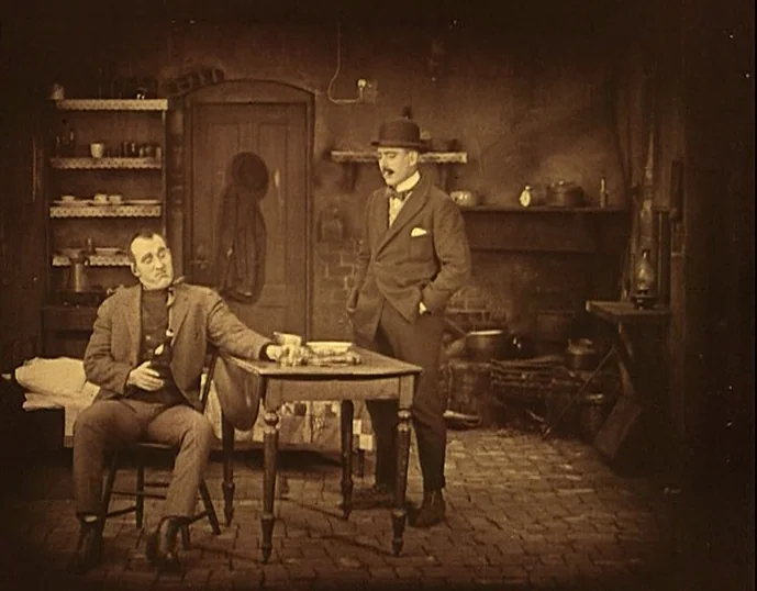 Broken Blossoms (1919) movie still: fullbody — A sparsely furnished room with brick flooring and a wooden table in the center. Two men interact in…