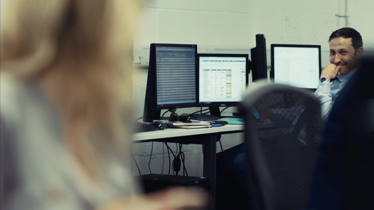 You Might Get Lost (2021) movie still: medium — A cluttered workstation with multiple computer monitors displays data sheets in a white-walled…