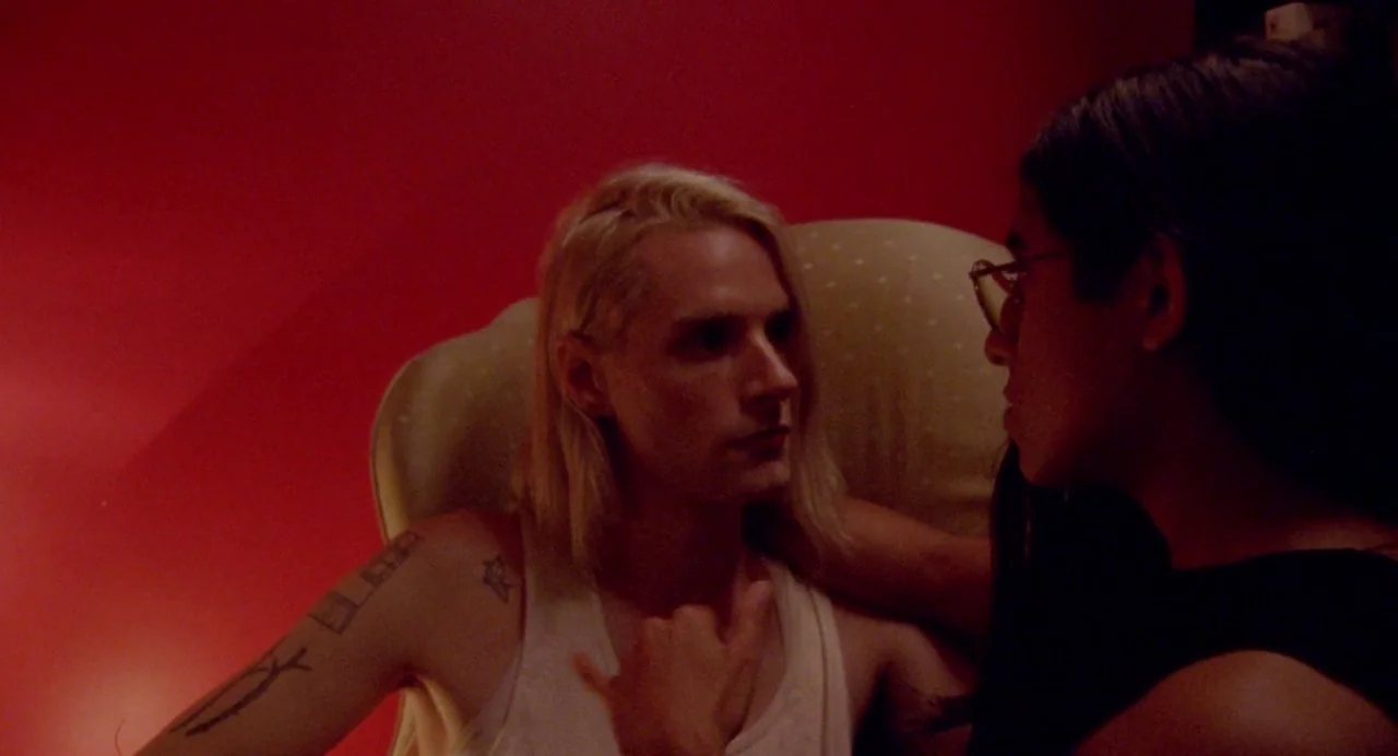 So Pretty (2019) movie still: medium — A dark, red-walled room features two individuals in close proximity during the night. A person with blonde…