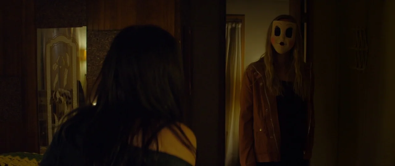 The Strangers: Prey at Night (2018) movie still: medium — A dimly lit room features wood-paneled walls and a figure wearing a plastic mask. The…
