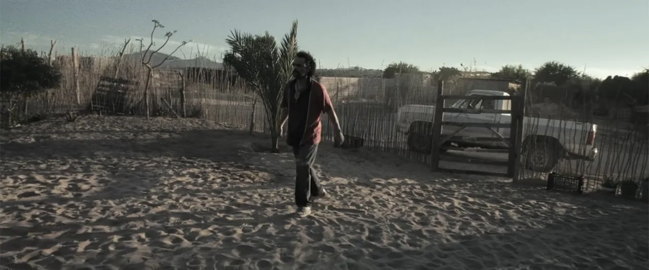At Dawn (2013) movie still: fullbody — A dry, sandy outdoor area features a wooden gate and a parked white pickup truck. A man walks across the sand…