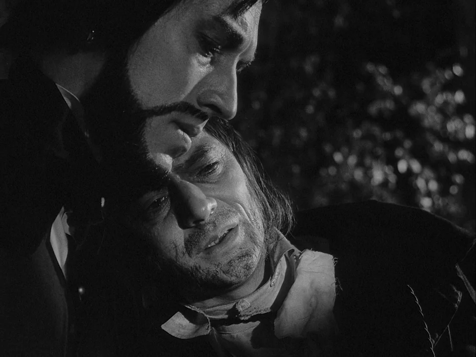 The Magician (1958) movie still: closeup — An outdoor setting at night with out-of-focus foliage in the dark background. One man leans his head onto…