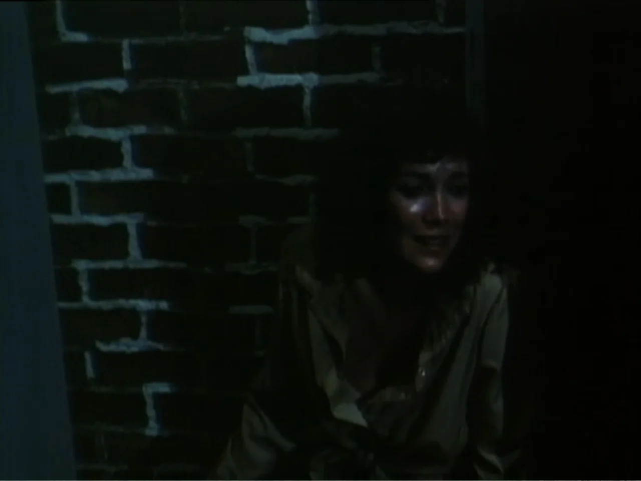 Curtains (1995) movie still: medium — A dark indoor space features a rough textured brick wall in the background. A woman in a tan shirt stands in…