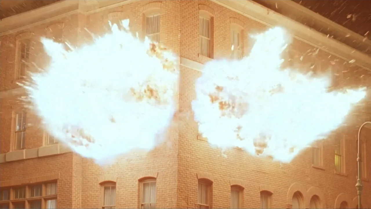 Entre las sombras (2025) movie still: wide — A historic multi-story brick building facade serves as the backdrop for two massive fireballs. Debris…