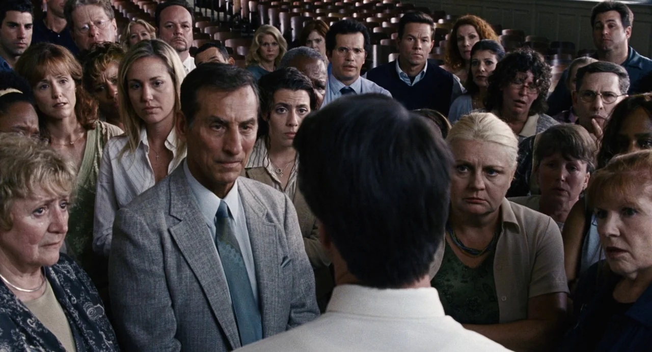 The Happening (2008) movie still: medium — An auditorium filled with a diverse crowd of adults in casual and business attire. The people are densely…