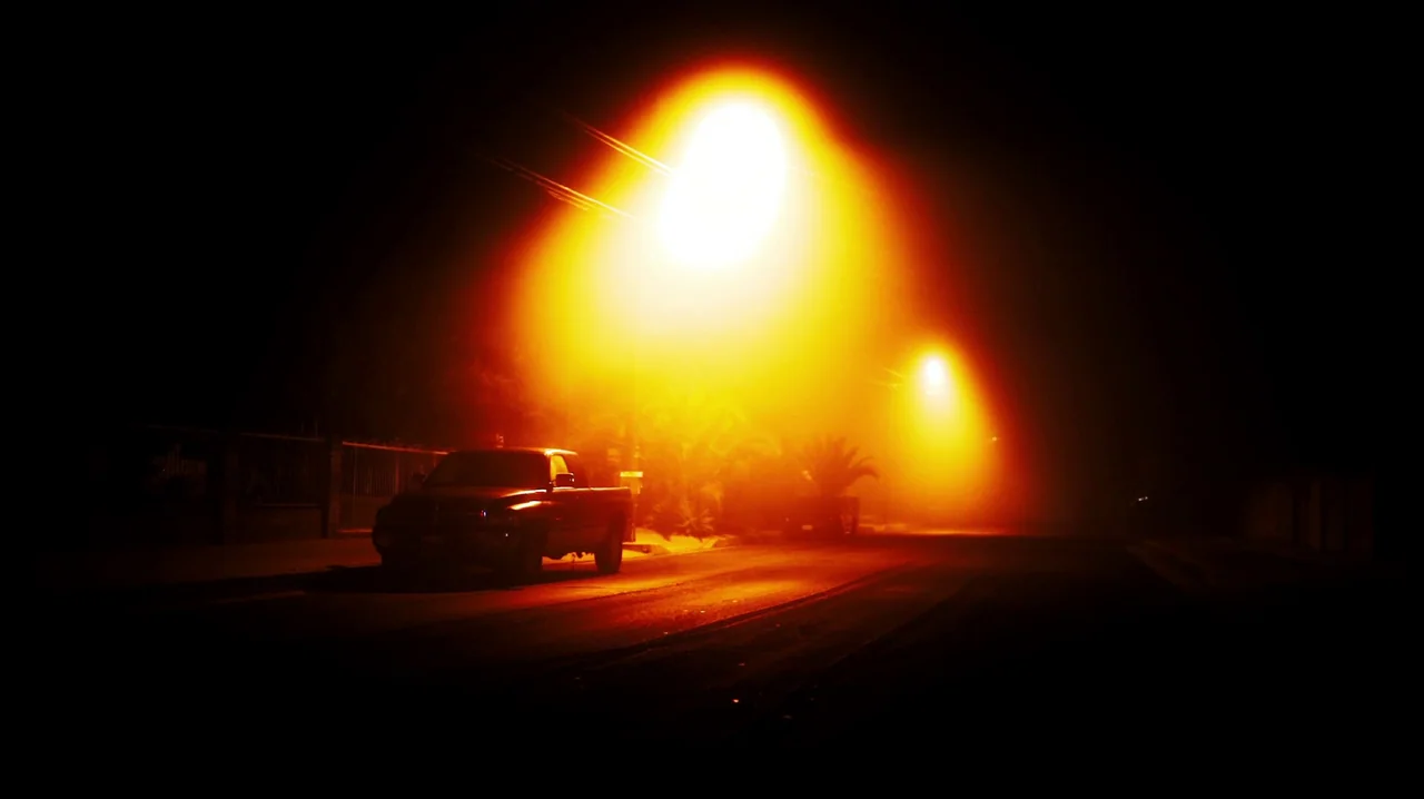 Carnage for Christmas (2024) movie still: wide — A quiet residential street is shrouded in thick night fog and orange streetlamp glare. A parked…