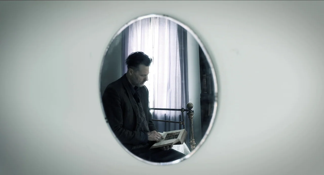 Where The Devil Roams (2023) movie still: medium — An oval-shaped mirror frame reveals a man sitting on a metal-framed bed. He is looking down at an…