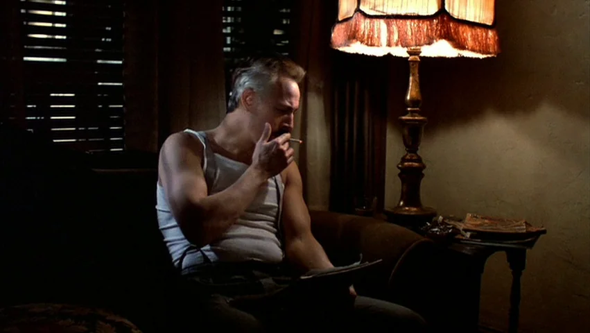 Hammett (1982) movie still: medium — A dimly lit room with heavy drapes covering the windows at night. A middle-aged man in a tank top sits by an…