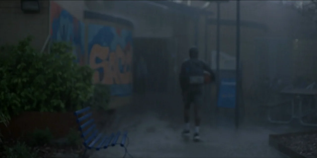 Bring Her Back (2025) movie still: wide — A school courtyard during a rainy night with graffiti on a tan wall. A young male student walks away from…