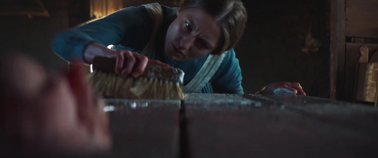 The Wind (2018) movie still: medium — A dark, dimly lit cabin interior features rough-hewn wooden surfaces and a rustic atmosphere. A young woman…