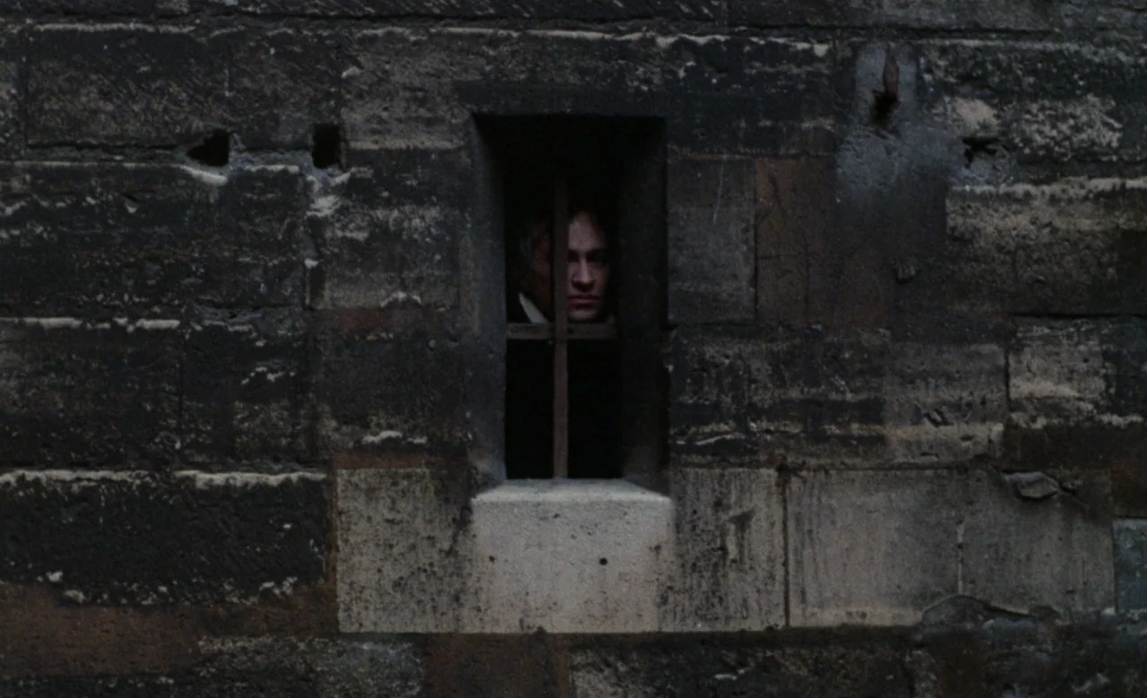 Blanche (1971) movie still: wide — A dark, weathered stone wall features a narrow vertical window frame with iron bars. A person's face appears in…