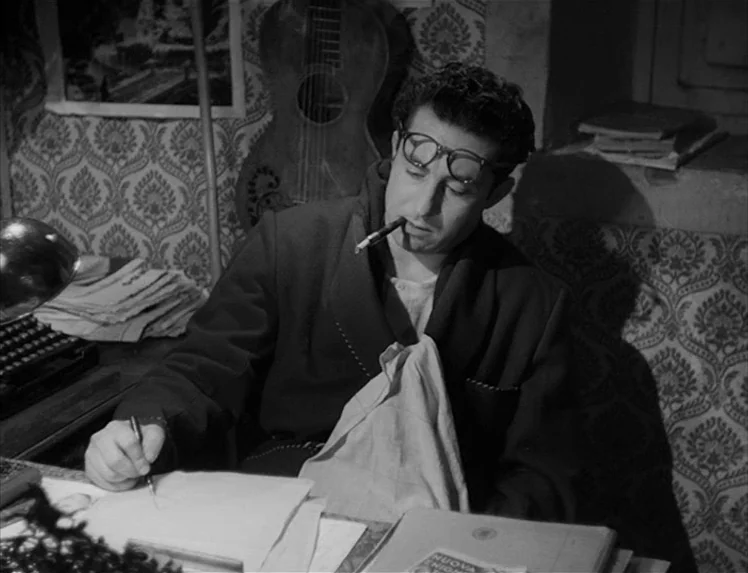 I Vitelloni (1953) movie still: medium — A cluttered workspace featuring patterned wallpaper and an acoustic guitar hanging behind the desk. A man…