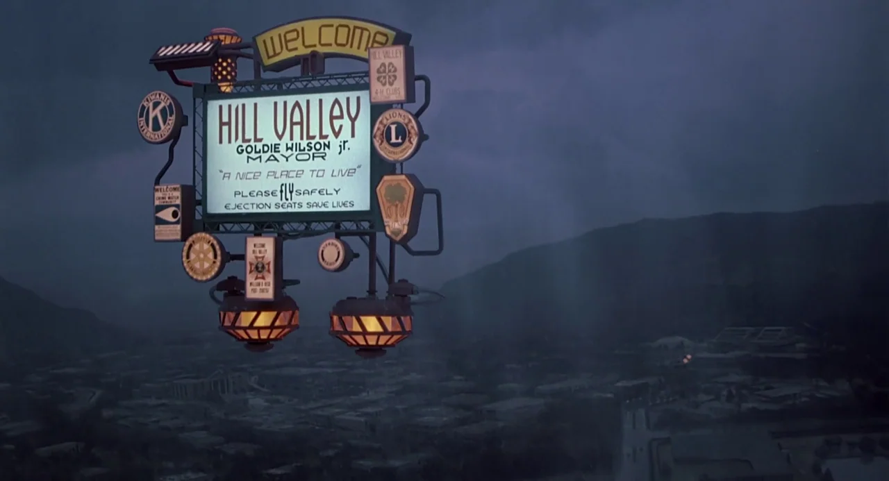 Back to the Future II (1989) movie still: establishing — A welcome sign for Hill Valley stands at the outskirts of a dark town. The sign features…