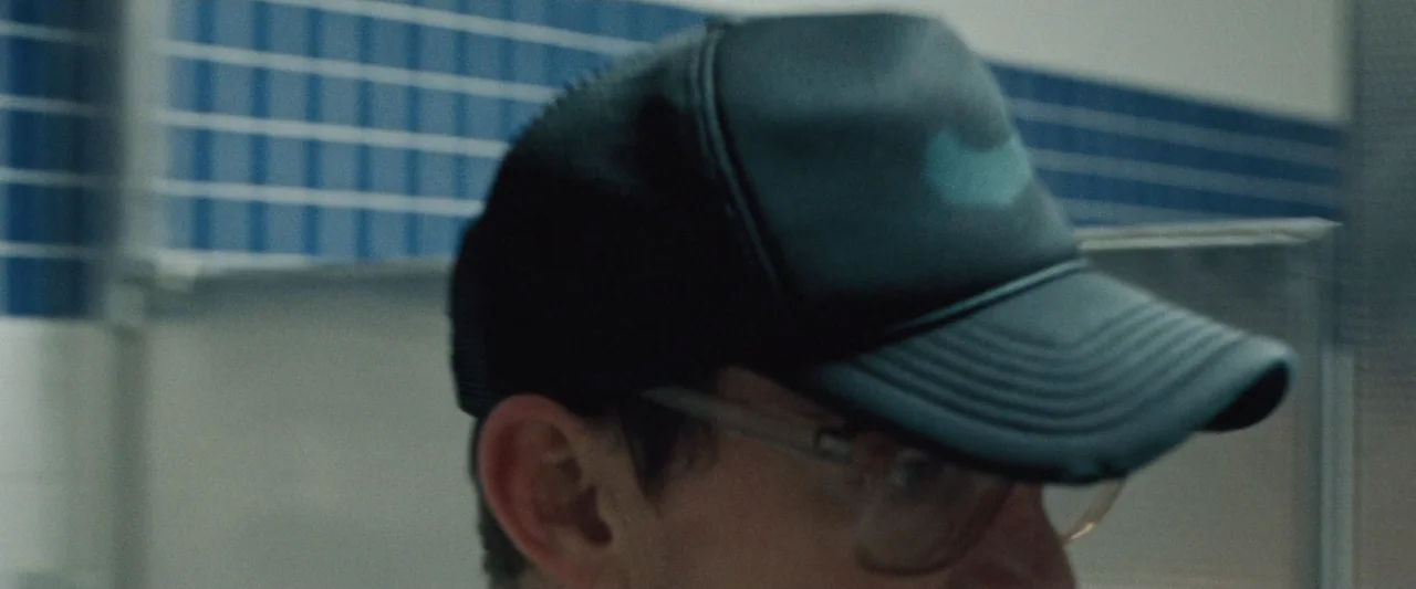 Roofman (2025) movie still: closeup — A public restroom interior with blue tiled walls and a grey partition wall. A man wearing a black baseball cap…