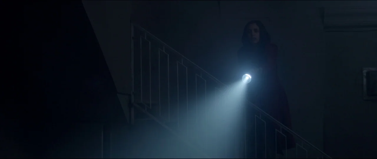 Under The Shadow (2016) movie still: medium — A dark staircase features a metal banister railing leading into deep shadows. A woman stands on the…