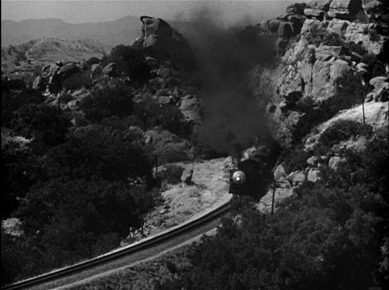 White Heat (1949) movie still: establishing — A steam train exits a rock tunnel surrounded by trees and rugged mountain slopes. The locomotive emits…