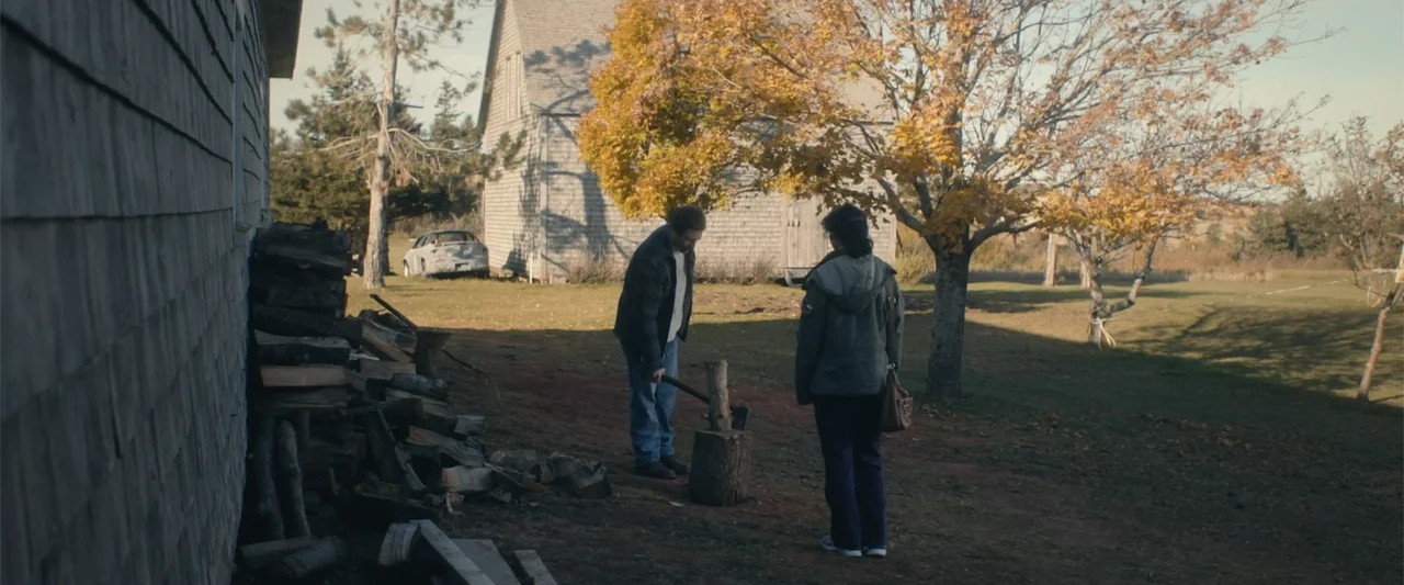 A Small Fortune (2021) movie still: wide — A rural yard features a wooden house and a large tree with autumn leaves. A man standing over a stump…