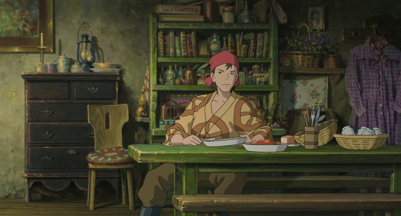 The Boy and The Heron (2023) movie still: medium — A cluttered, rustic room with a painted green table and shelves full of objects. A man with a red…