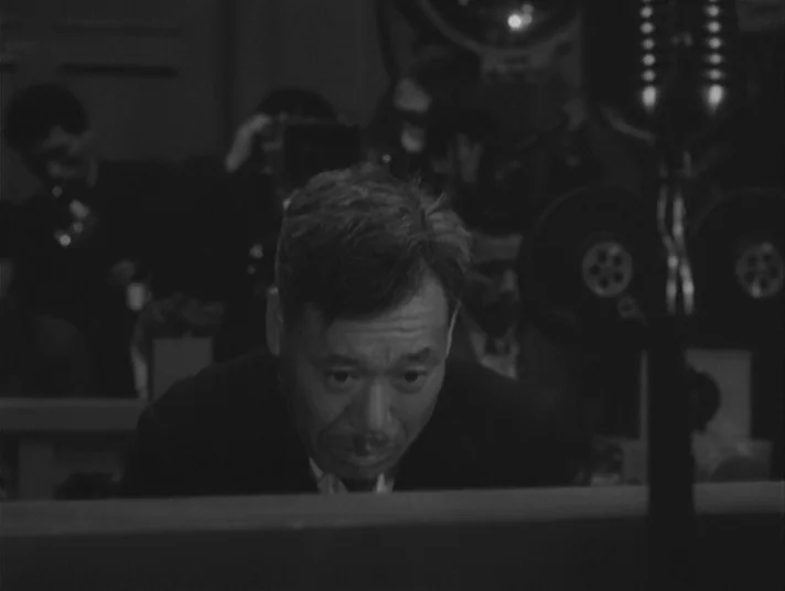 Scandal (1950) movie still: medium — A dimly lit room containing audio equipment including a prominent vertical microphone. An Asian man in a dark…