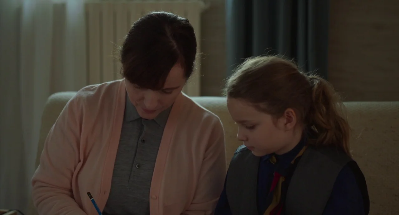 Petite Maman (2021) movie still: medium — A woman and a young girl sit together in a room with neutral walls. They are focused on a task involving…