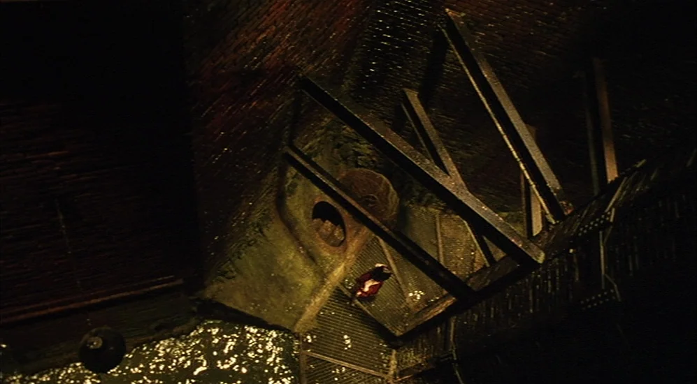 The City Of Lost Children movie still: wide — A dark brick building features a network of industrial iron staircases and grating. A single person…