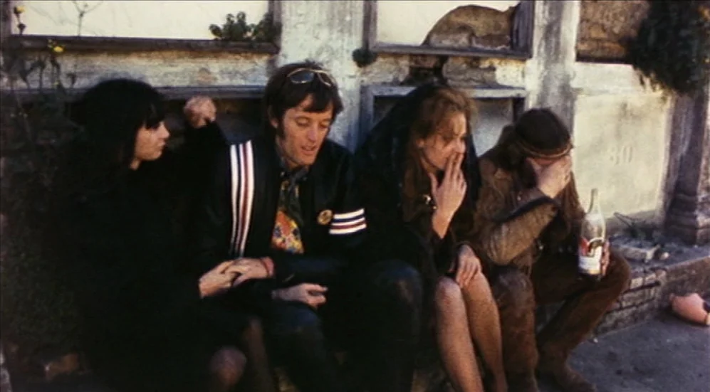 Easy Rider (1969) movie still: medium — Four young people sit on the ground against a weathered stone wall with recessed panels. They are positioned…