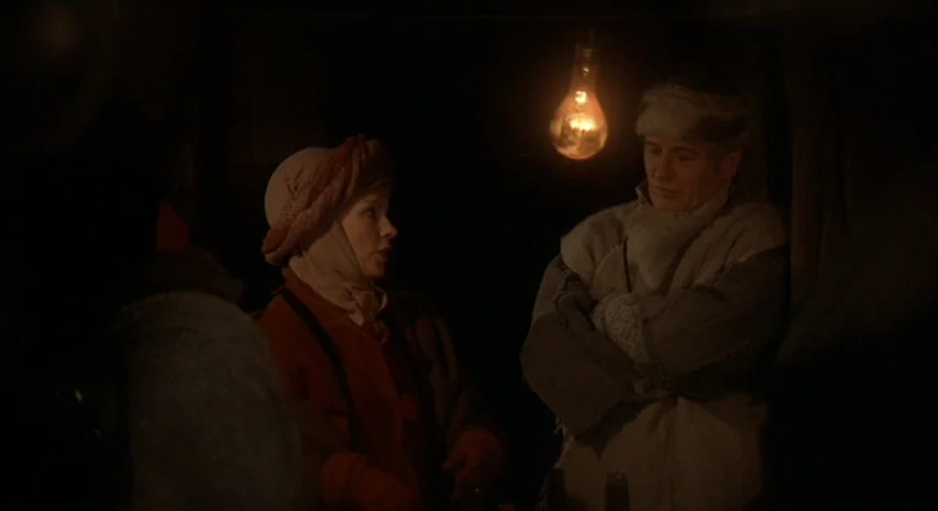 Quintet (1979) movie still: medium — Two individuals sit in a dark room illuminated only by a single bare bulb. They wear thick winter clothing and…