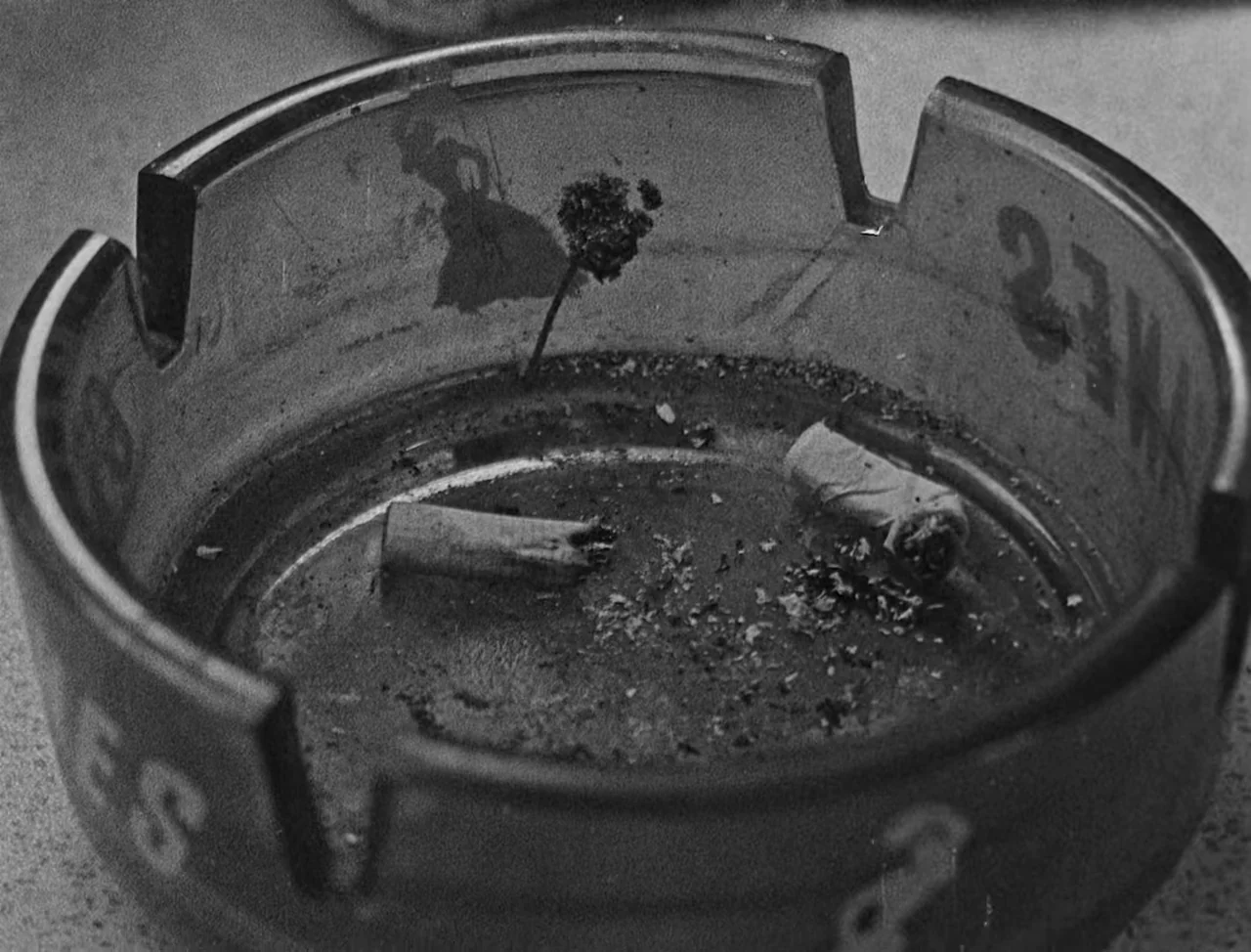 Bait (2019) movie still: closeup — A dark glass ashtray sits on a light-colored surface with specks of debris. Two extinguished cigarette butts lie…