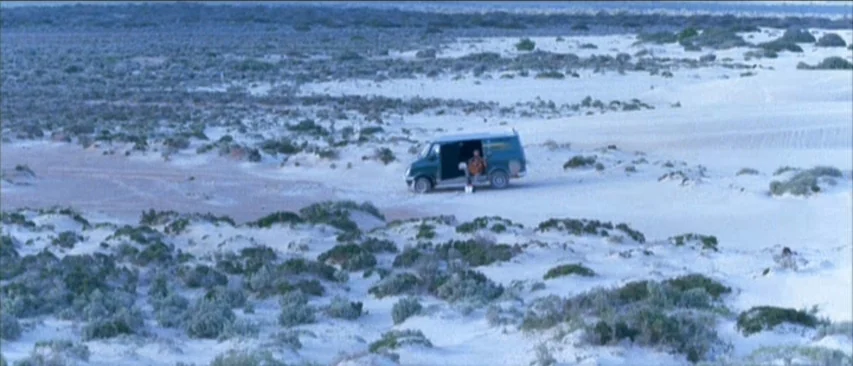 Roadgames (1981) movie still: wide — A vast expanse of white sand dunes under an overcast, light blue sky. A single small dark green van is parked…