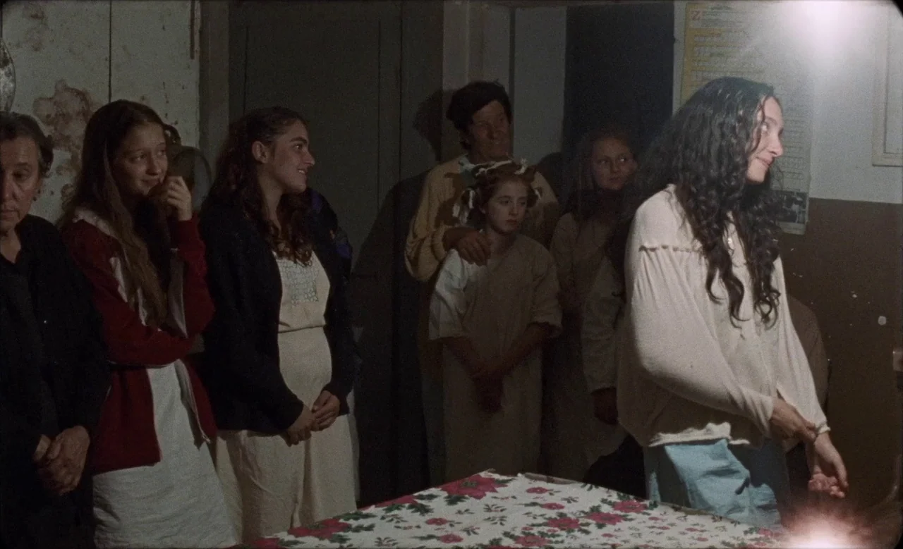 Happy as Lazzaro (2018) movie still: medium — A dimly lit room with multiple people standing together in a gathering. The woman in the foreground…
