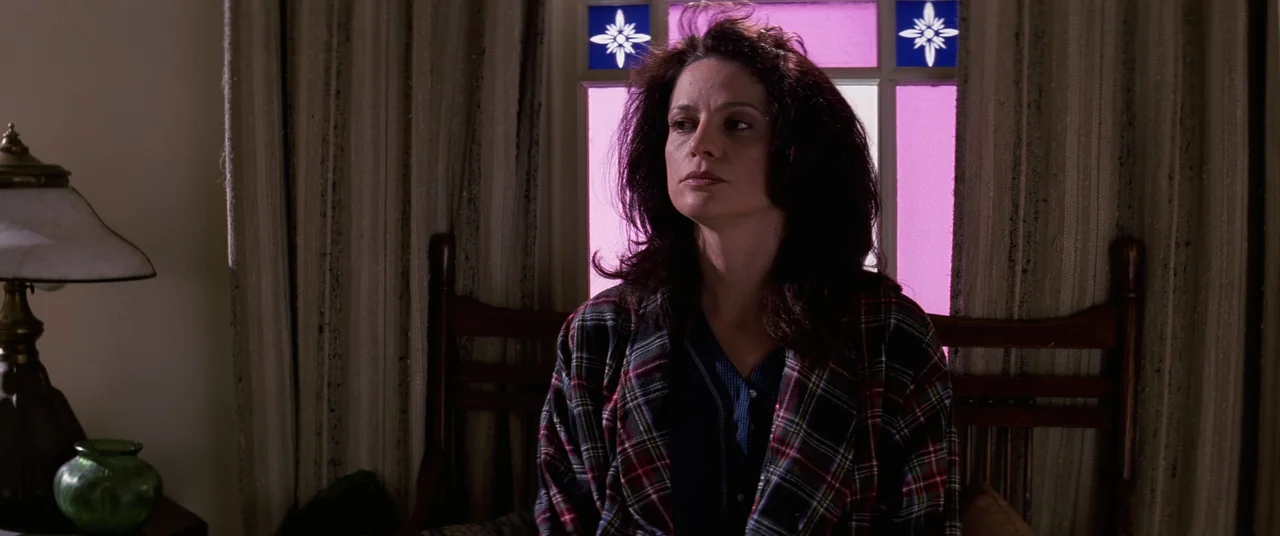 Hackers (1995) movie still: medium — A woman sits in a bedroom before a window with pink glass detail. The room contains a wooden headboard and a…
