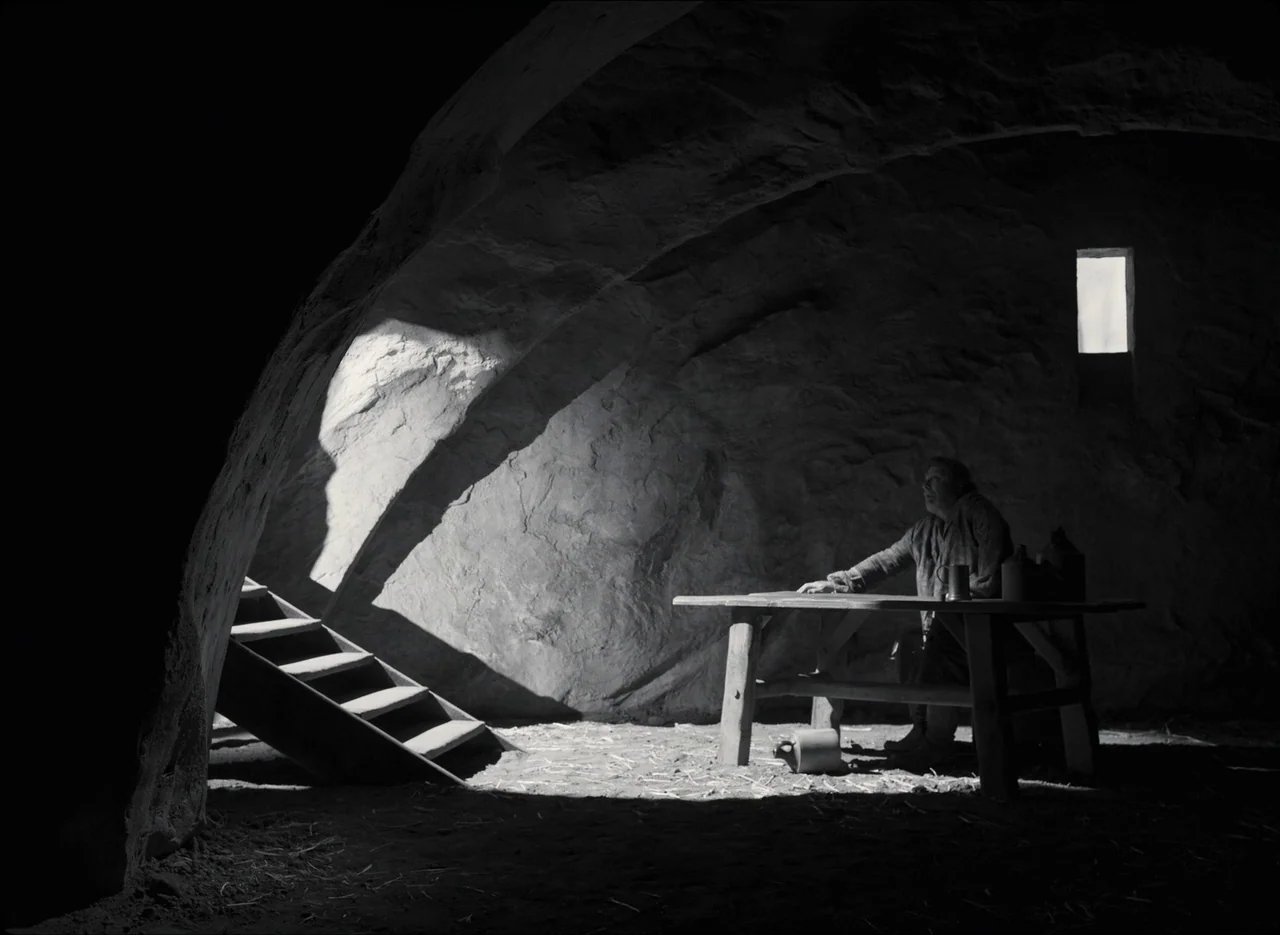 The Tragedy of Macbeth (2021) movie still: wide — A dark, vaulted stone space with an uneven floor and a wooden staircase. A single man sits at a…