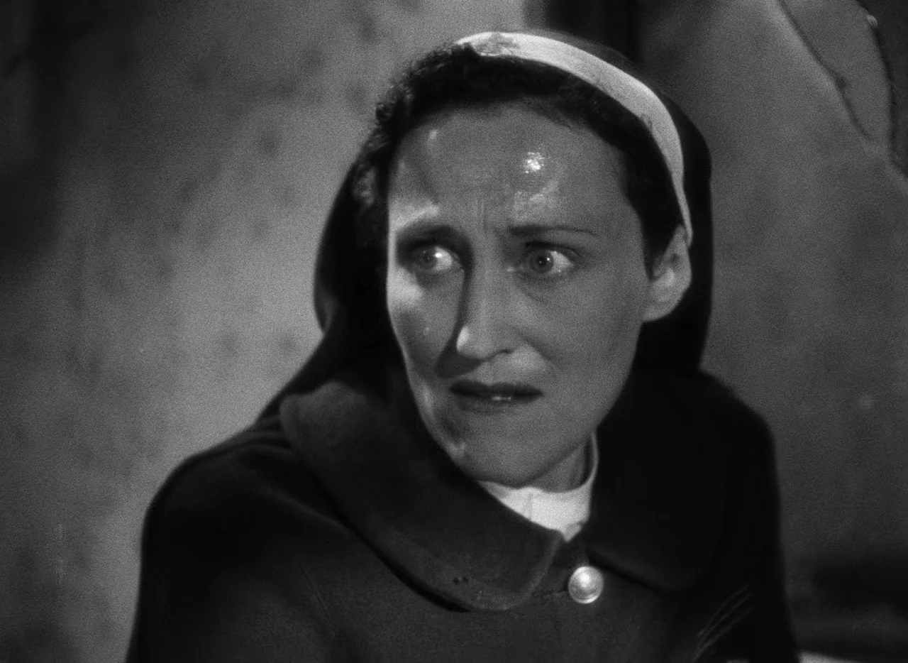 Le Corbeau (1943) movie still: closeup — A dimly lit interior space with a plain wall background. A nun in a traditional habit glances sharply…