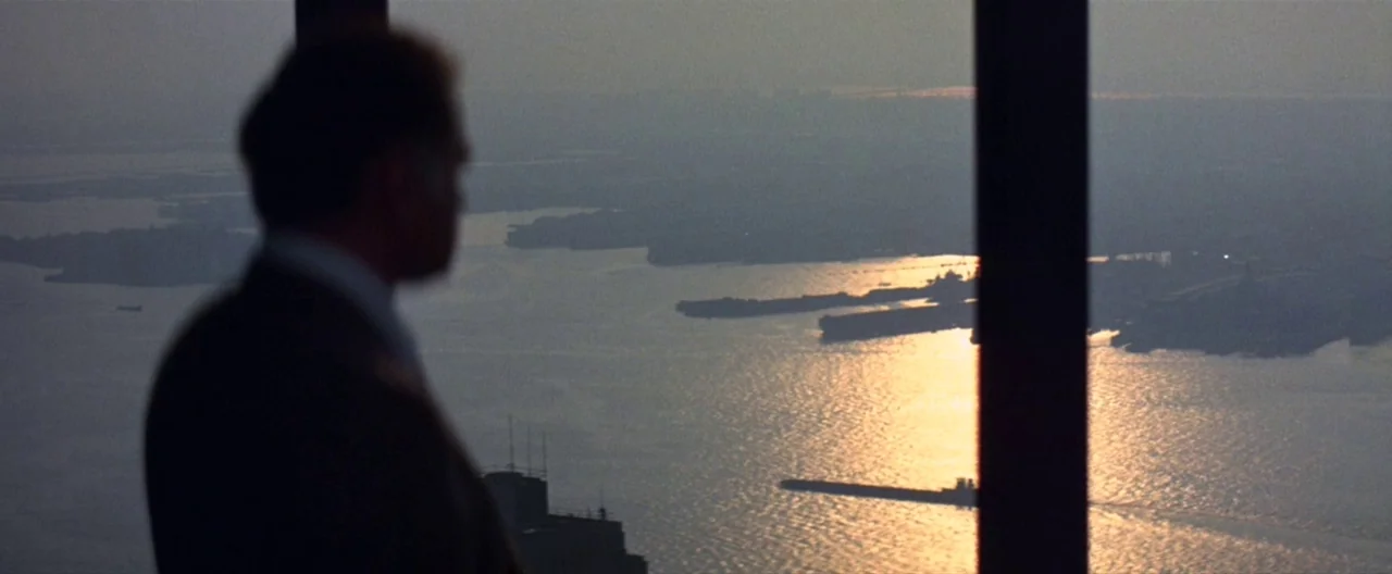 Klute (1971) movie still: medium — A high-rise office overlooks a large body of water during the late afternoon. A man in a dark suit stands…