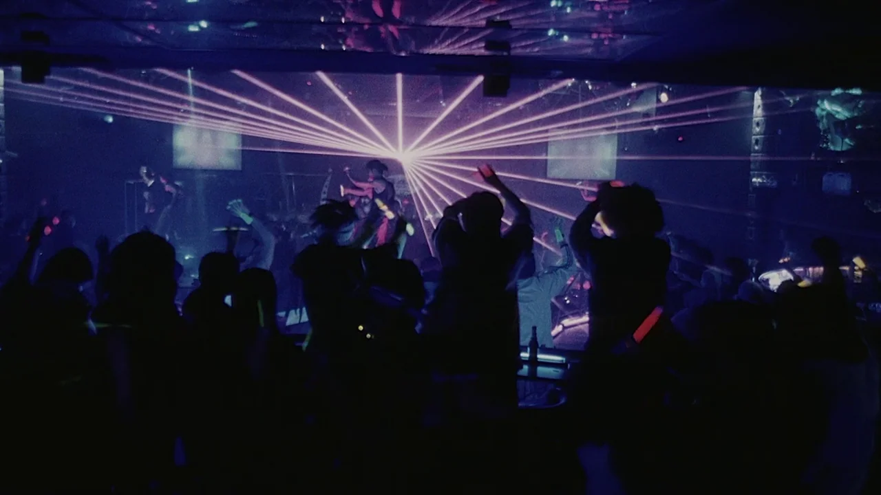 Visible Secret (2001) movie still: wide — A darkened interior space features a crowd of people in silhouette against pink laser light. The room is…