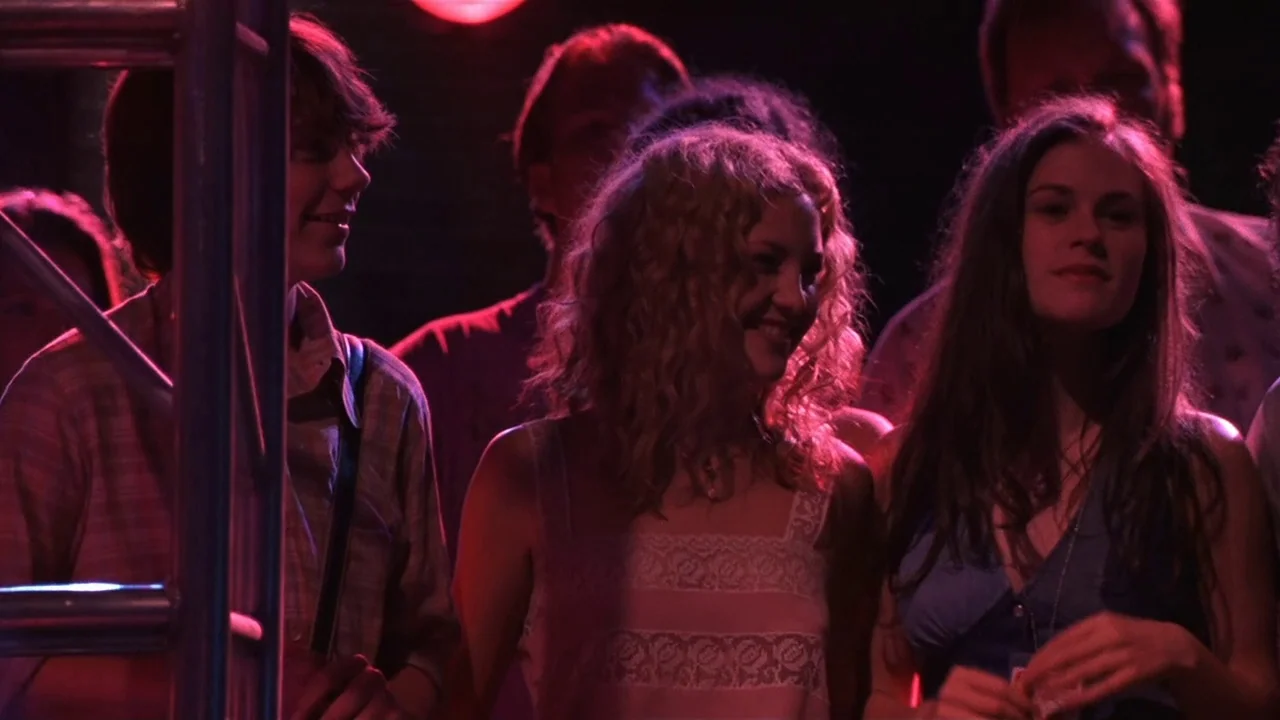 Almost Famous (2000) movie still: medium — A dark concert venue is illuminated by vibrant red stage lights. Three young people stand together in a…