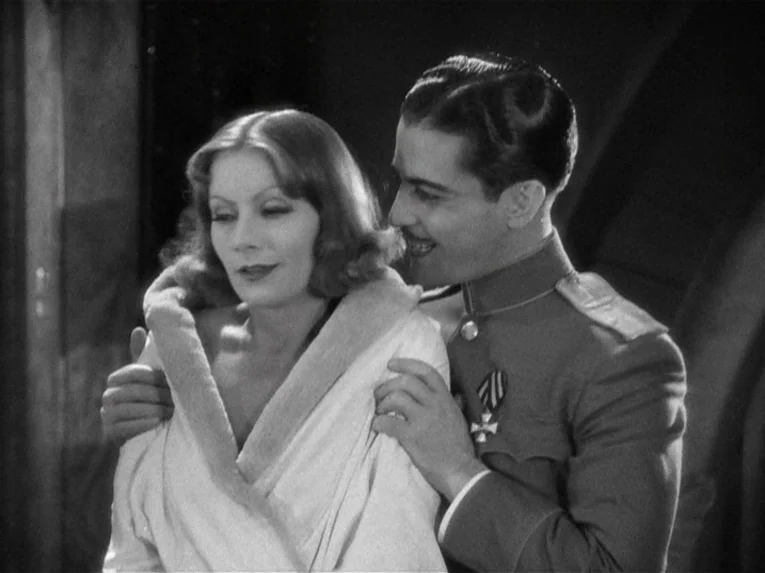 Mata Hari (1931) movie still: medium — An interior space with a man in military uniform standing behind a woman. The woman wears a dressing gown…