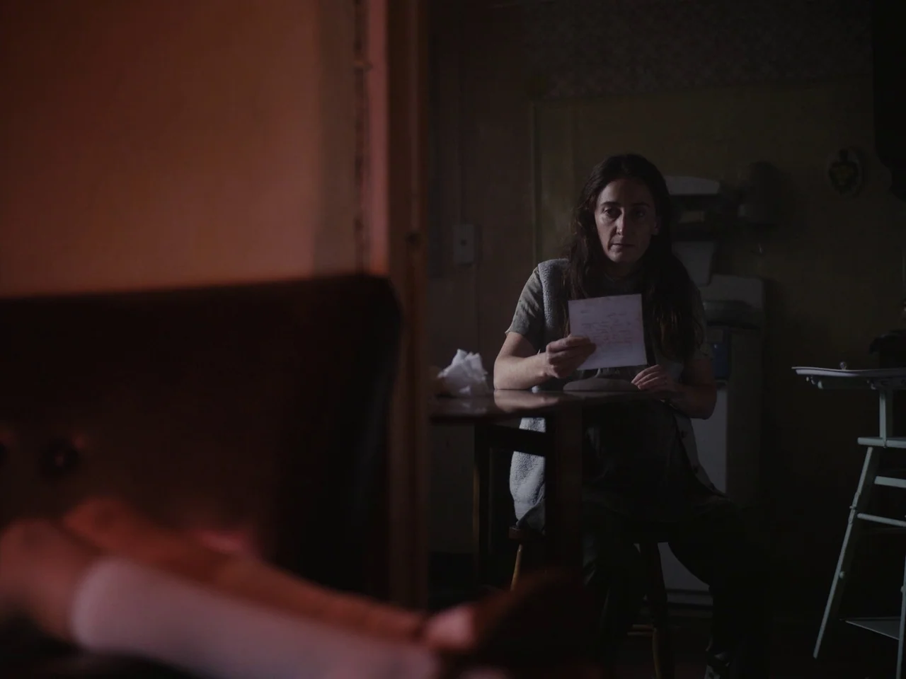 The Quiet Girl (2022) movie still: medium — A dimly lit room contains a woman seated at a wooden table. The woman looks toward the foreground while…