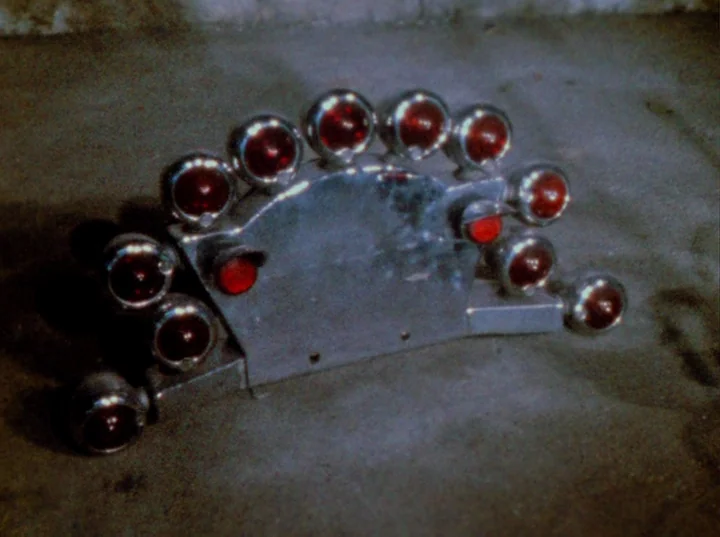 Scorpio Rising (1963) movie still: closeup — A metallic semi-circular object with multiple red glass indicators sits on a concrete floor. The object…
